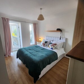 A bedroom with a bed, a bedside table, a lamp, and a window with curtains. - Modern Bright Riverside Stay (London)