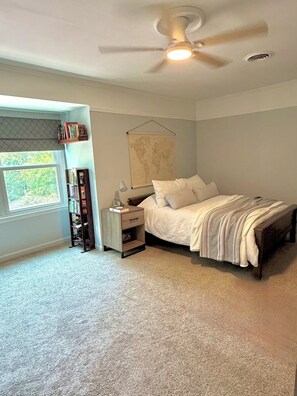 A bedroom with a bed, a nightstand, a lamp, a bookshelf, a ceiling fan, and a window with a view of greenery. - 5BR Private Aiken Home on 2 Acres - 30 Min to Augusta National - Masters Week (Aiken)