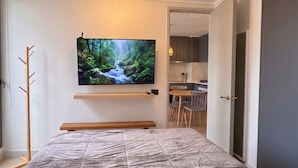 A modern bedroom with a bed, a mounted TV displaying a nature scene, a coat rack, and an open door leading to a dining area. - Santiago Metro Ecuador - Domus Travels (Santiago)