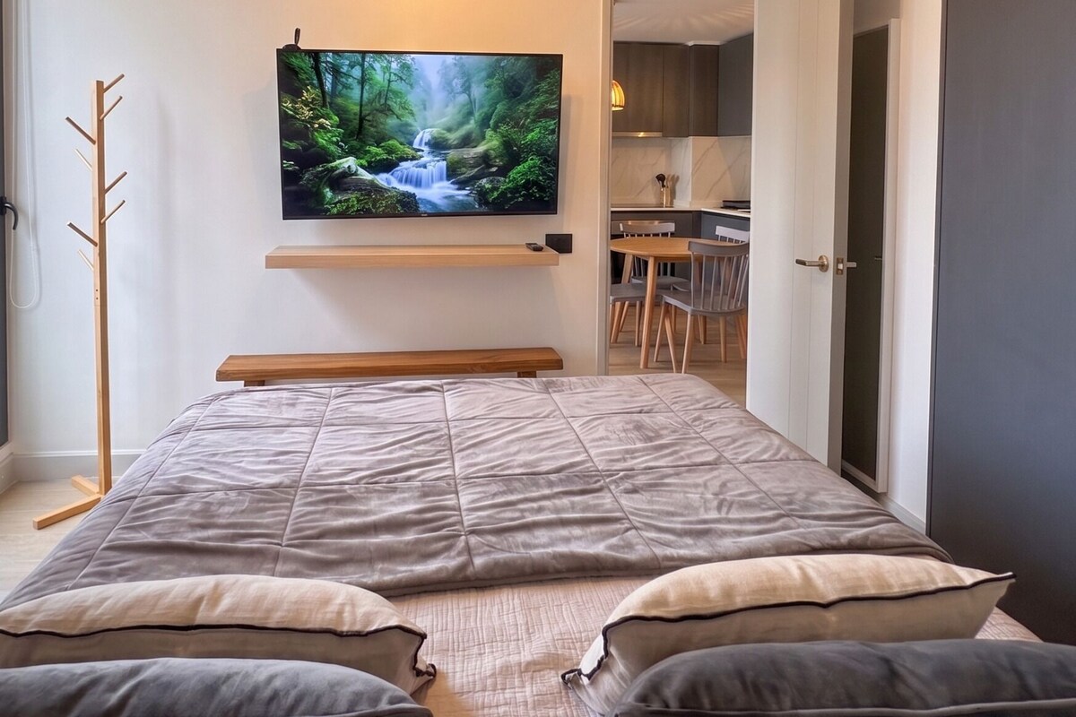 A modern bedroom with a bed, a mounted TV displaying a nature scene, a coat rack, and an open door leading to a dining area.