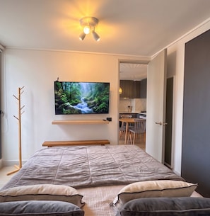 A modern bedroom with a bed, a mounted TV displaying a nature scene, a coat rack, and an open door leading to a dining area. - Santiago Metro Ecuador - Domus Travels (Santiago)