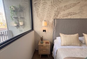 Ein Schlafzimmer mit einem Bett, einem Nachttisch mit Lampe, einem Fenster mit Blick auf Gebäude und einer Wand mit Blattmuster.