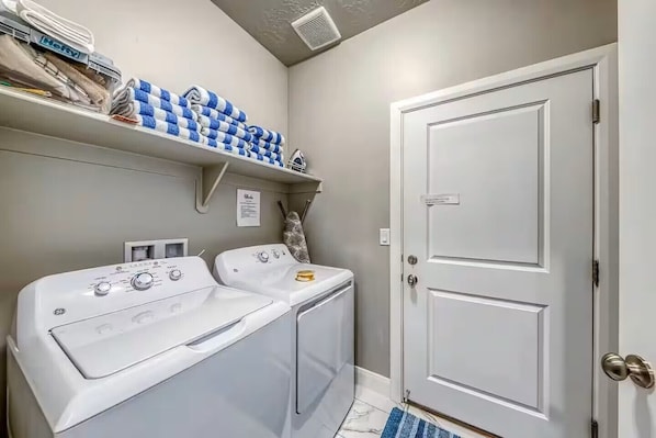 A laundry room with two front-loading washers and dryers, a shelf with folded towels, and a closed door.