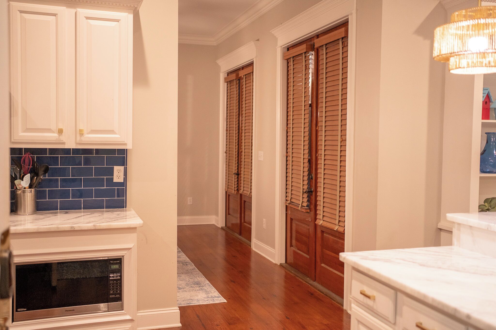A hallway with wooden doors, a kitchen area with white cabinets, and a marble countertop.