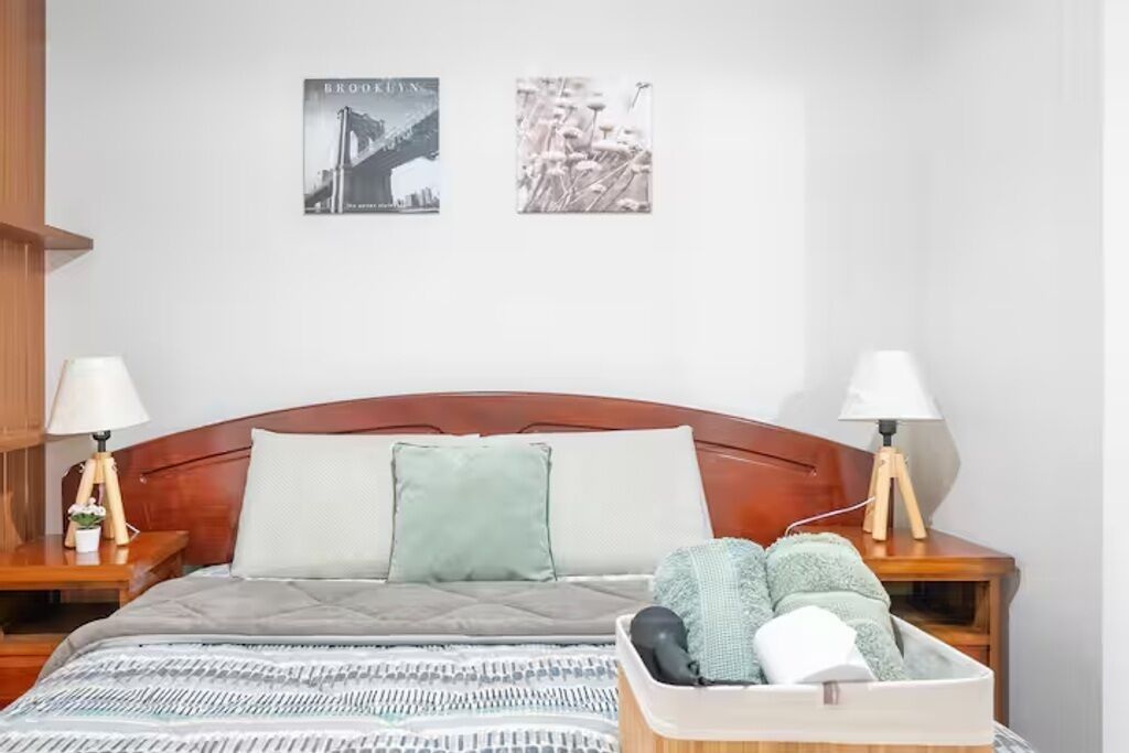 A neatly made bed with a grey quilt, two bedside lamps, a small bedside table with a lamp, and a wall-mounted picture frame.