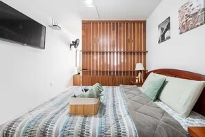Un dormitorio con una cama, cabecera de madera, televisor montado en la pared y una lámpara de noche.