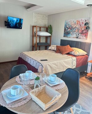 Ein Hotelzimmer mit einem Bett, einem an der Wand befestigten Fernseher, einem Gemälde und einem Essbereich mit Tisch und Stühlen.