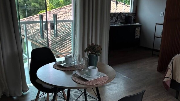 A dining area with a round table, two chairs, a vase with flowers, and a view of a balcony with a tiled roof. - Guest house 700 meters from Moçambique beach and 500 meters from the state park. (Florianópolis)