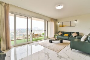 Un salón amplio con un sofá verde, una mesa de centro y vistas a un paisaje urbano a través de amplios ventanales correderas.