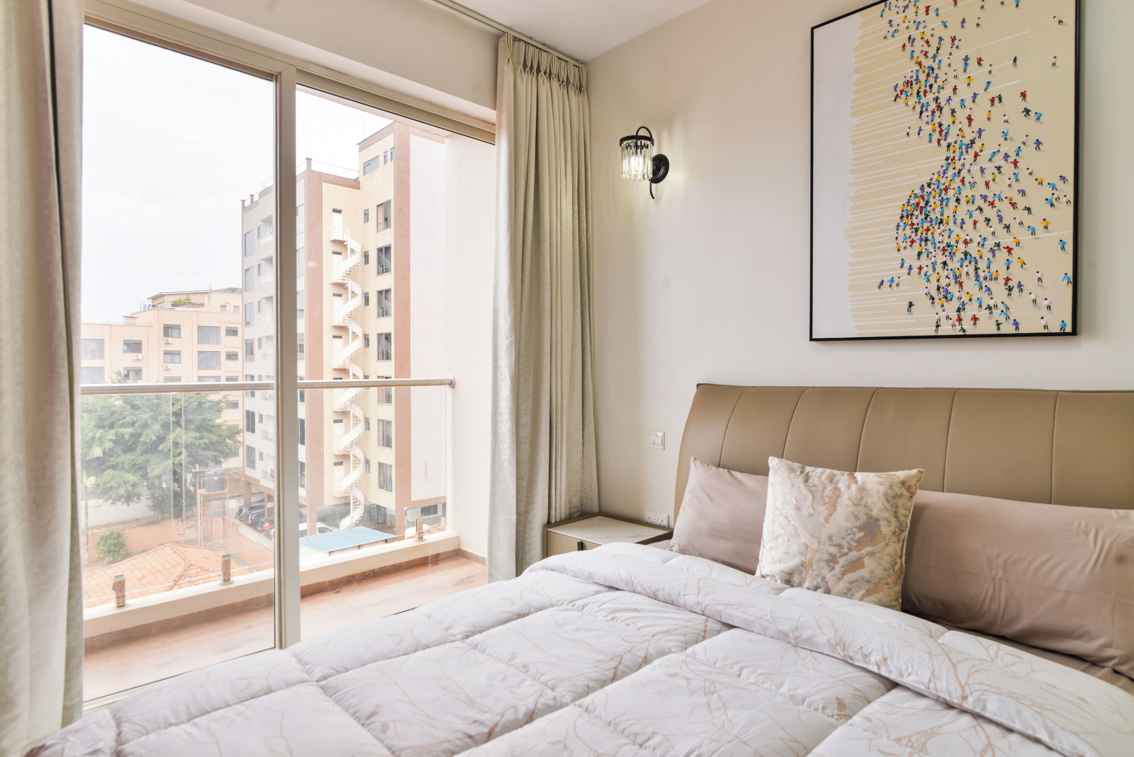 Habitación de hotel con una cama grande, vistas de edificios a través de una ventana y una lámpara fijada a la pared.