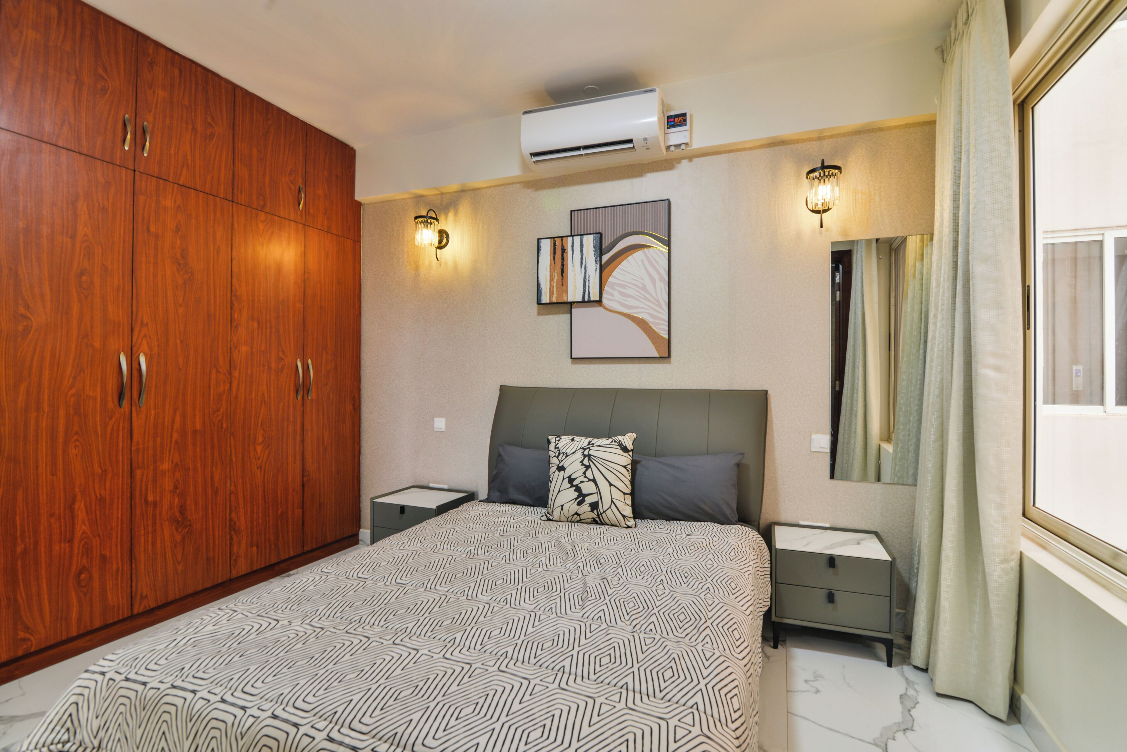 Un dormitorio con un armario grande, una cama con ropa de cama estampada, una mesita de noche, un aire acondicionado instalado en la pared y una lámpara de noche.