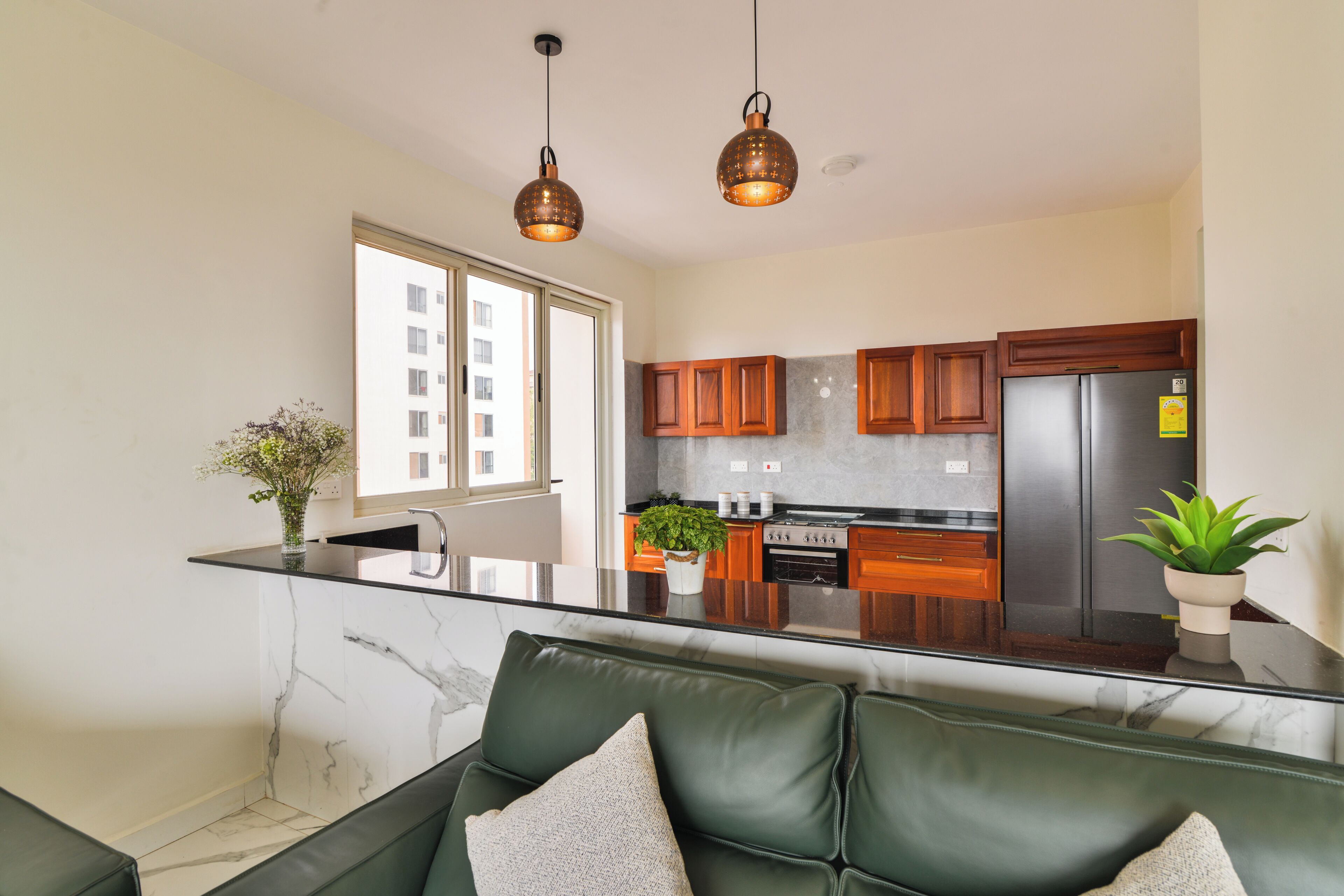 A modern kitchen with a marble countertop, wooden cabinets, a stainless steel refrigerator, and pendant lighting.