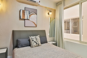 Habitación de hotel con cama, dos almohadas, mesita de noche, lámpara de pared, ventana con cortinas y una obra de arte en la pared.