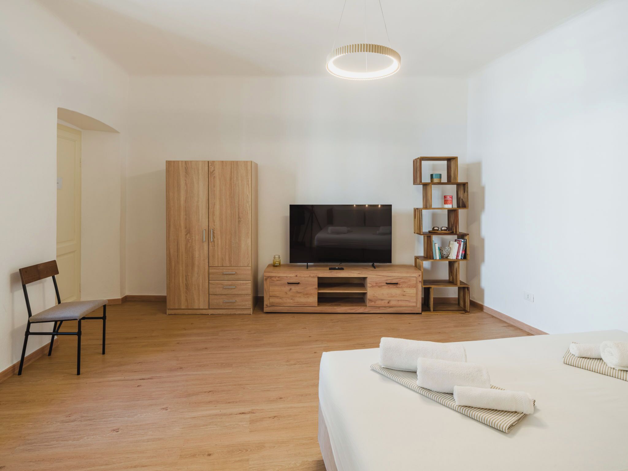 A hotel room with a wooden floor, a bed with white linens, a TV on a wooden stand, a wooden wardrobe, a wooden chair, and a wooden bookshelf.