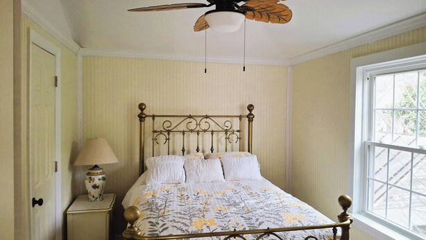 A bedroom with a bed, a nightstand with a lamp, a ceiling fan, and a window.