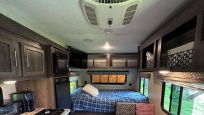 A compact RV interior with a kitchenette, a bed with a window, and a small seating area. - MINI Camper -fully equipped stationary (Champlain)