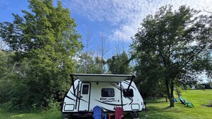 A white RV with a small porch area, two chairs, and a grassy field with trees in the background. - MINI Camper -fully equipped stationary (Champlain)