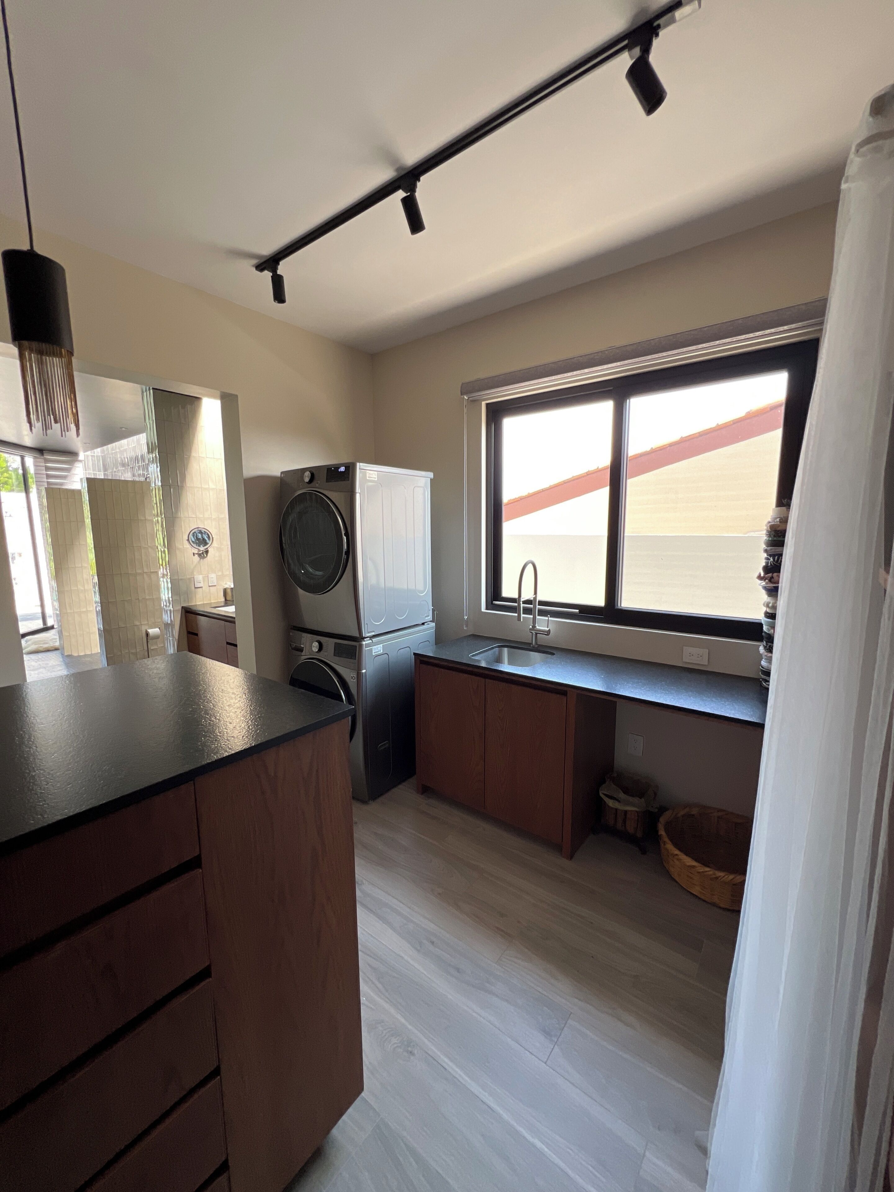 A modern kitchen with a built-in washer and dryer, wooden cabinets, a large window with a view, and a hanging light fixture.