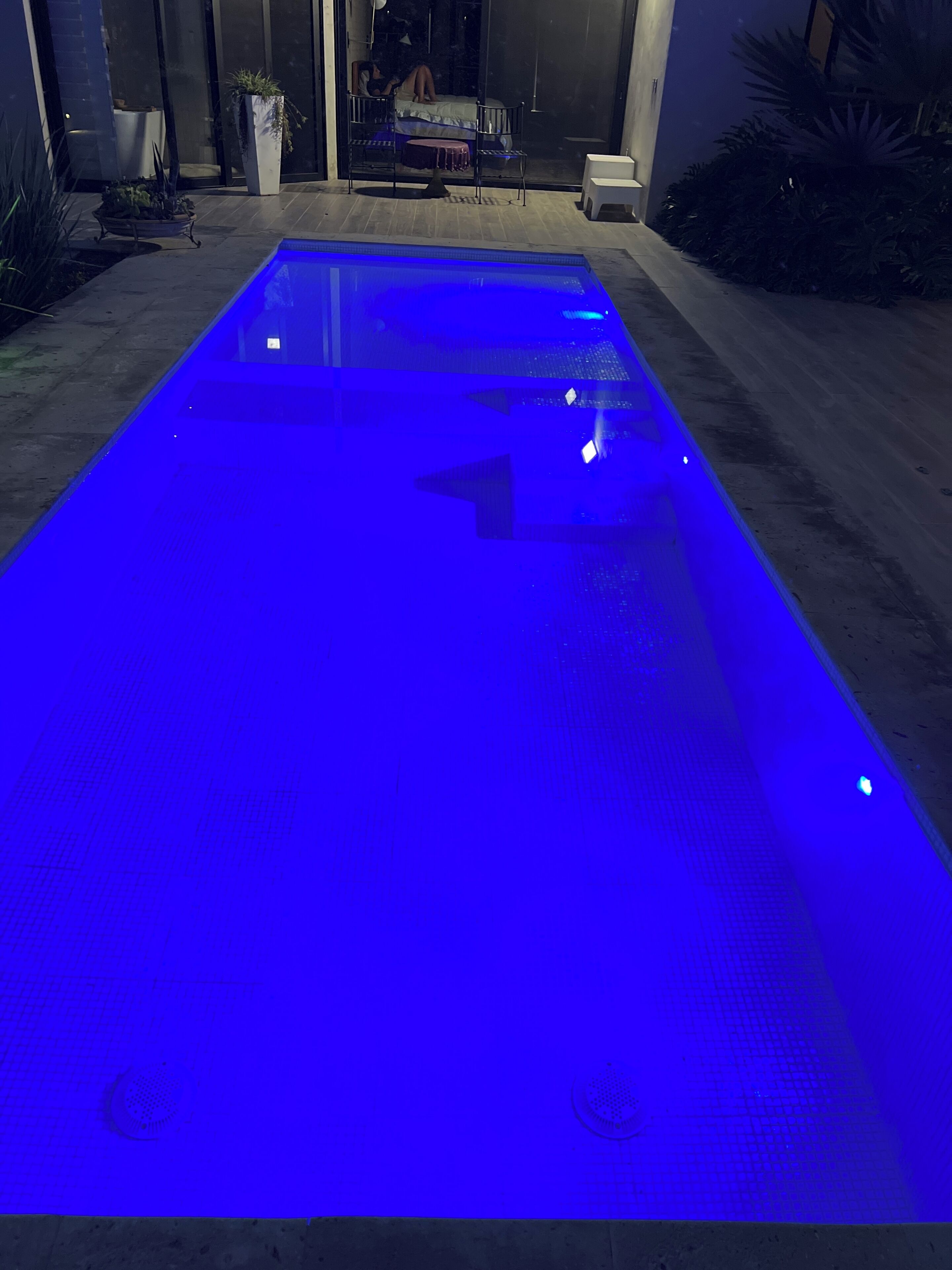 A swimming pool with a blue illuminated surface, surrounded by a patio area with seating and plants.