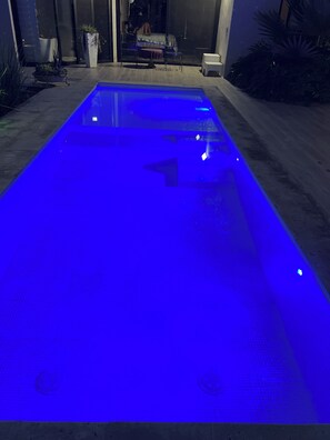 Une piscine dont la surface est bleue et illuminée, entourée d’un patio aménagé avec des sièges et des plantes.