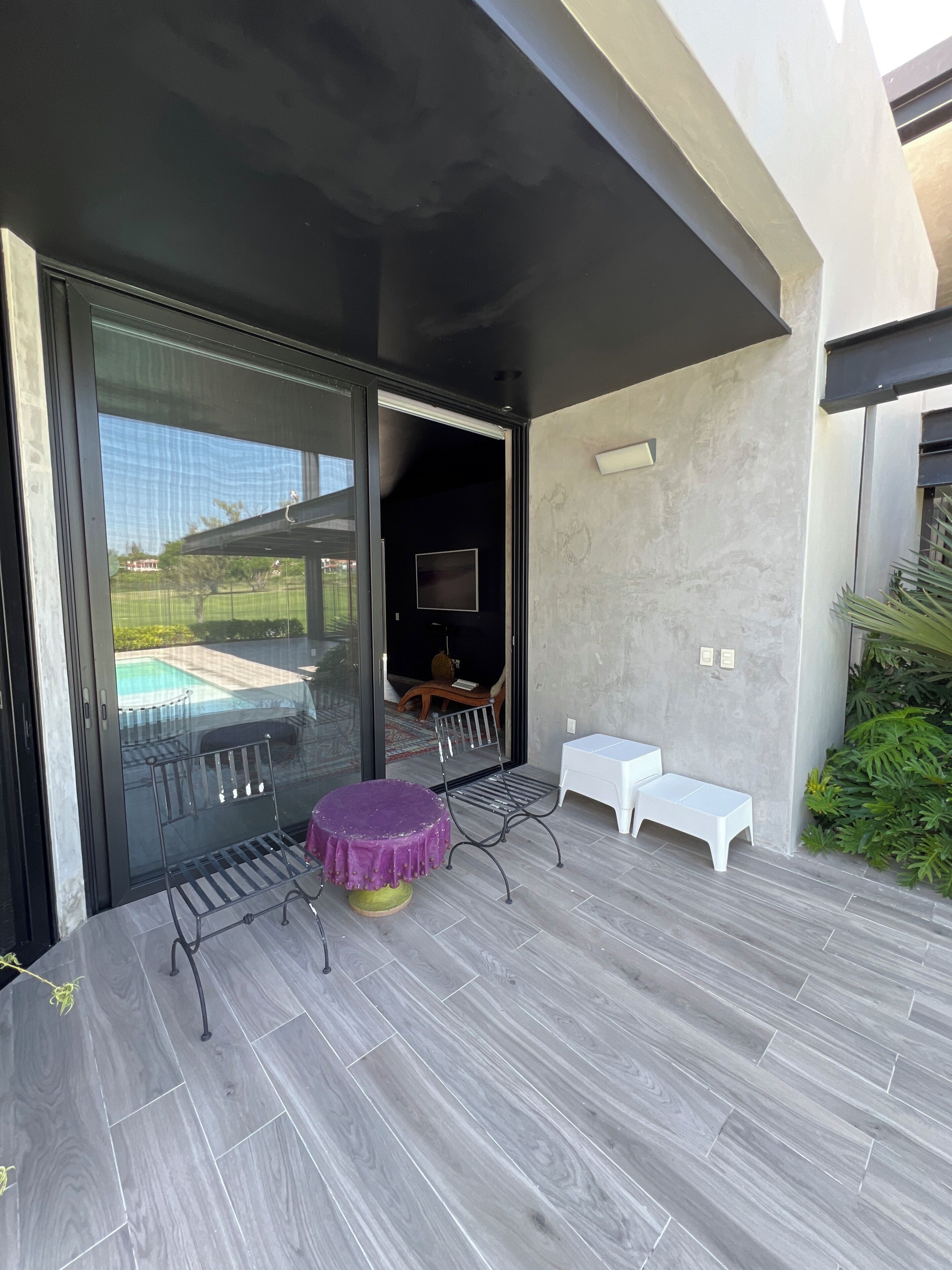 A modern outdoor patio with a glass door leading to a pool, a purple round table, a metal bench, and a small white table.