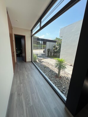 A corridor with a large window leading to an outdoor area with a gravel path, a cactus, and a parked car.