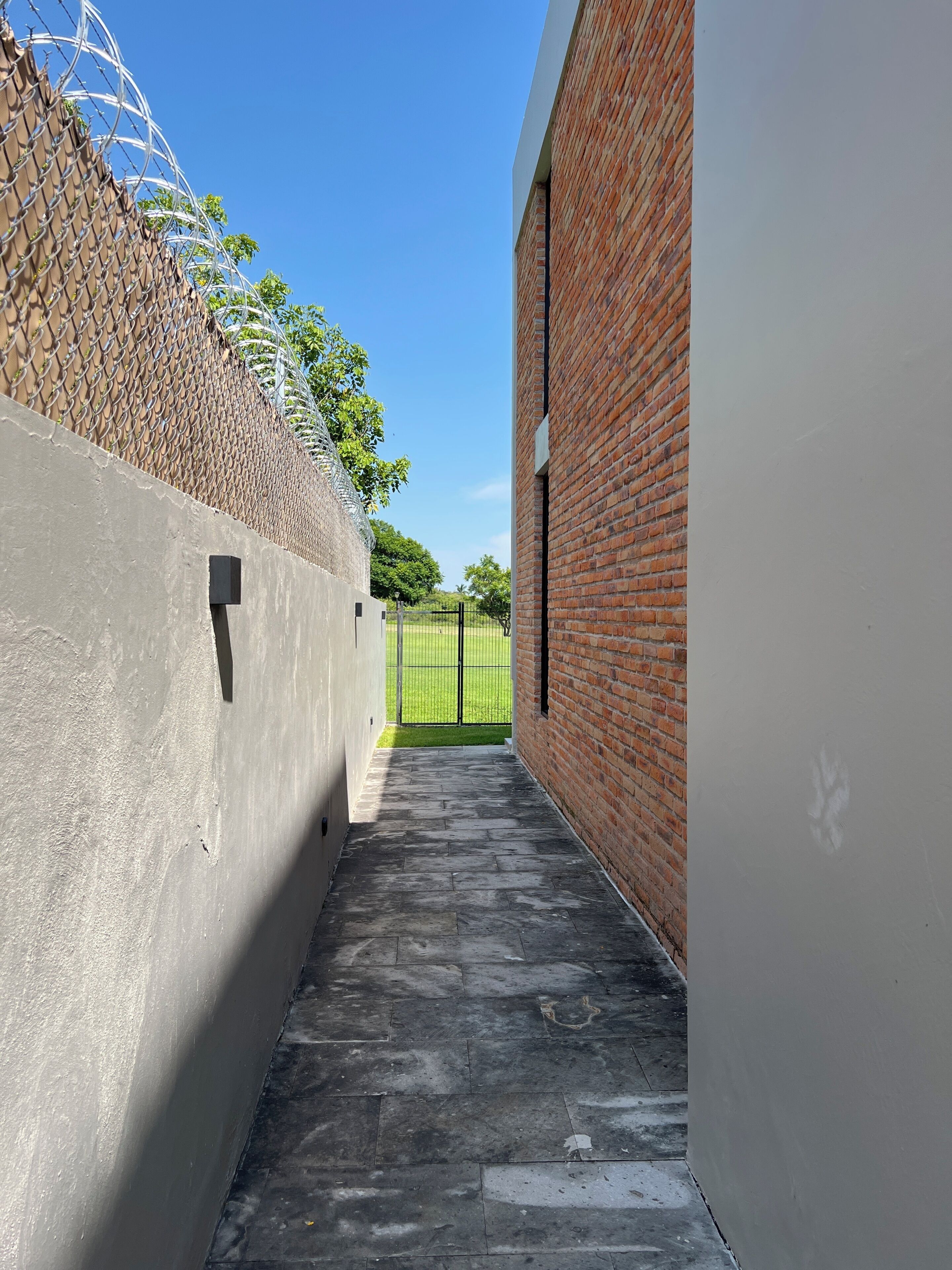 A narrow concrete pathway flanked by a brick wall and a chain-link fence, leading to a gated area with a grassy field visible in the distance.
