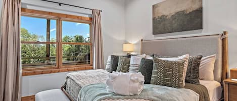 A bedroom with a large bed, a window with curtains, a bedside lamp, and a painting on the wall.