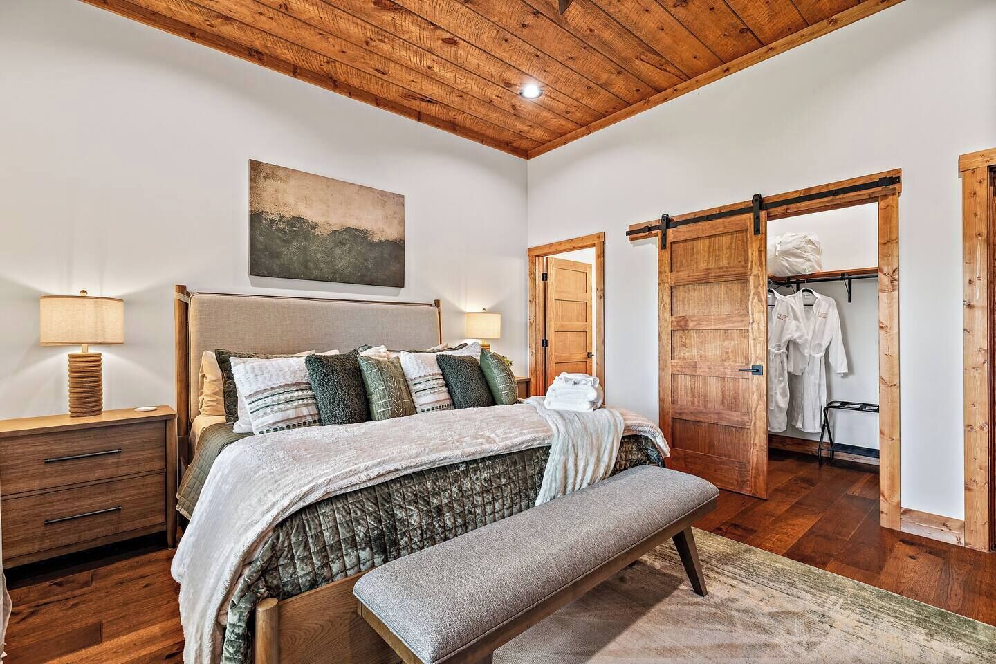 A bedroom with a wooden ceiling, a bed with a bench, a nightstand, a bedside table, and a wooden door leading to a bathroom with a towel rack.