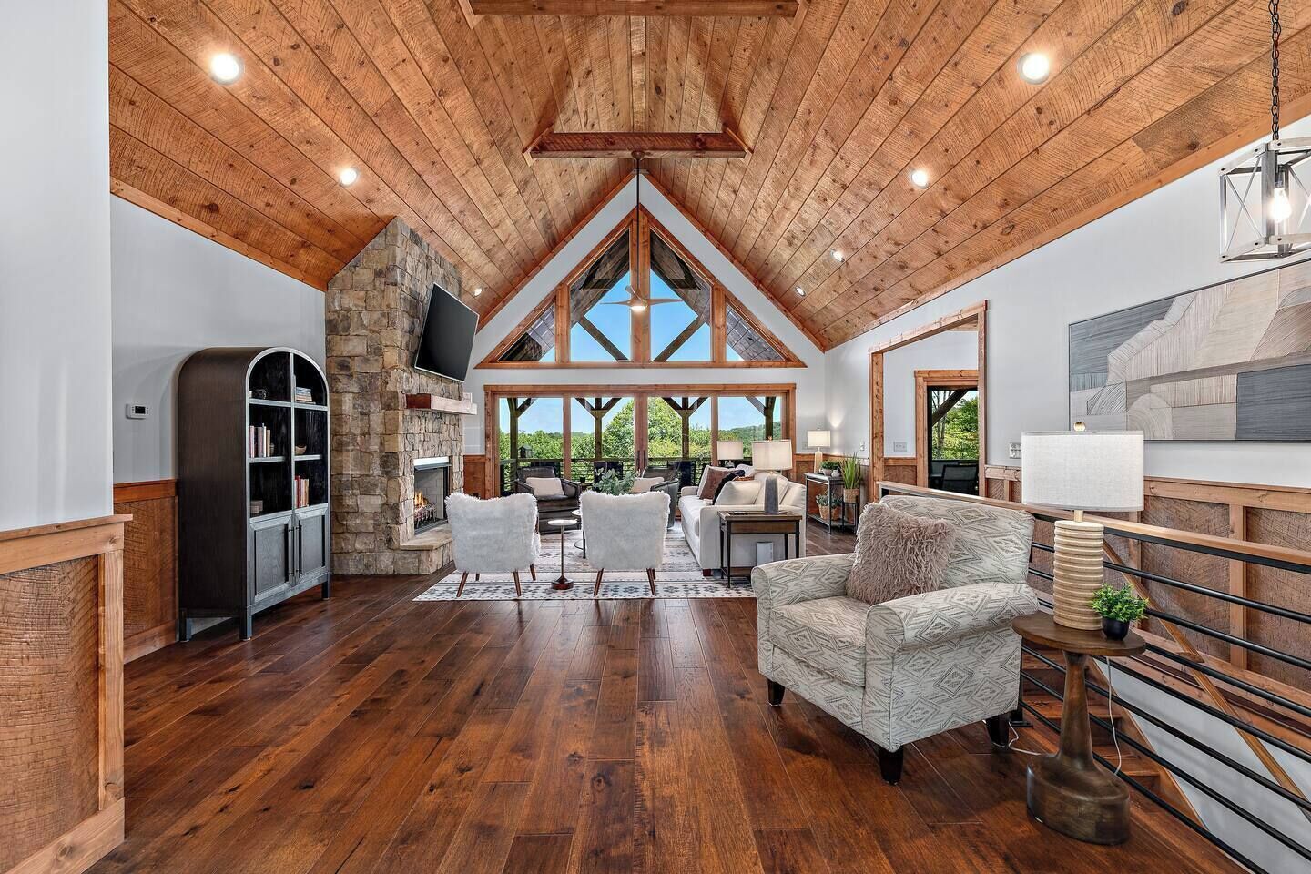 A spacious living area with a stone fireplace, wooden ceiling beams, and large windows offering a view of greenery.