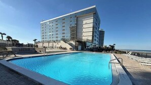 Una piscina con superficie de agua azul cristalina, rodeada por un patio empedrado. Al fondo, un gran edificio de hotel con varios pisos y fachada de vidrio.