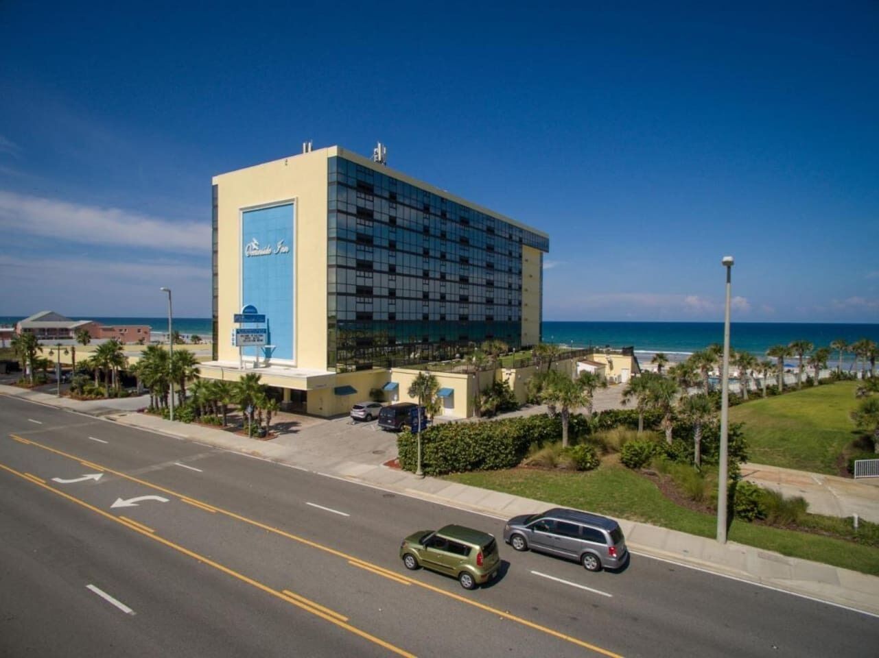 A hotel with a large glass facade, located near a beach with palm trees and a clear sky.