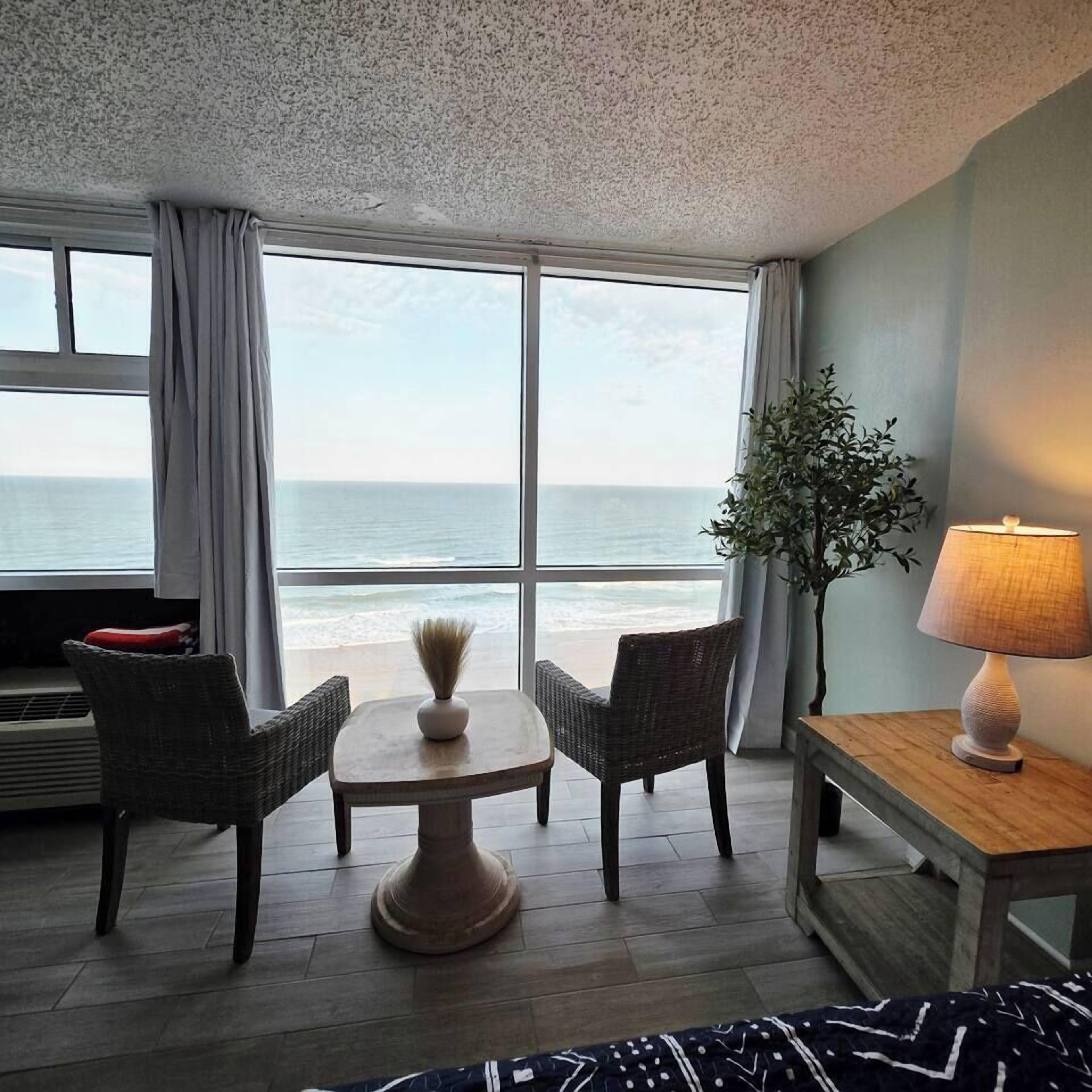 A room with a view of the ocean, a round table with chairs, a lamp, and a potted plant.