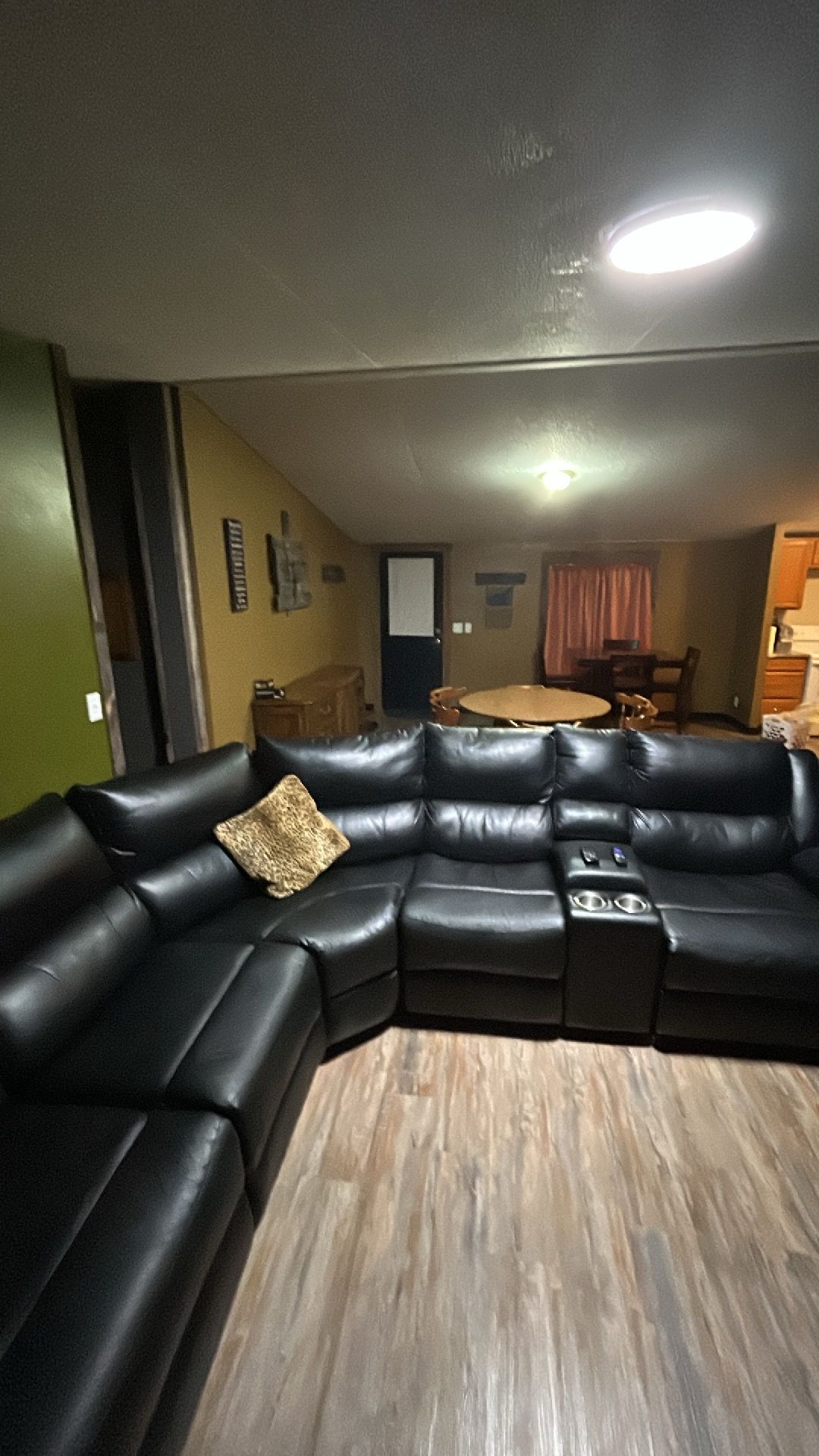 Una sala de estar con un sofá seccional de cuero negro, piso de madera, una mesa de comedor con sillas y una zona de cocina al fondo.