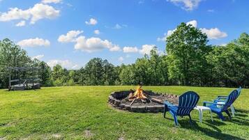 A circular fire pit with chairs and a table, surrounded by a grassy field and trees.