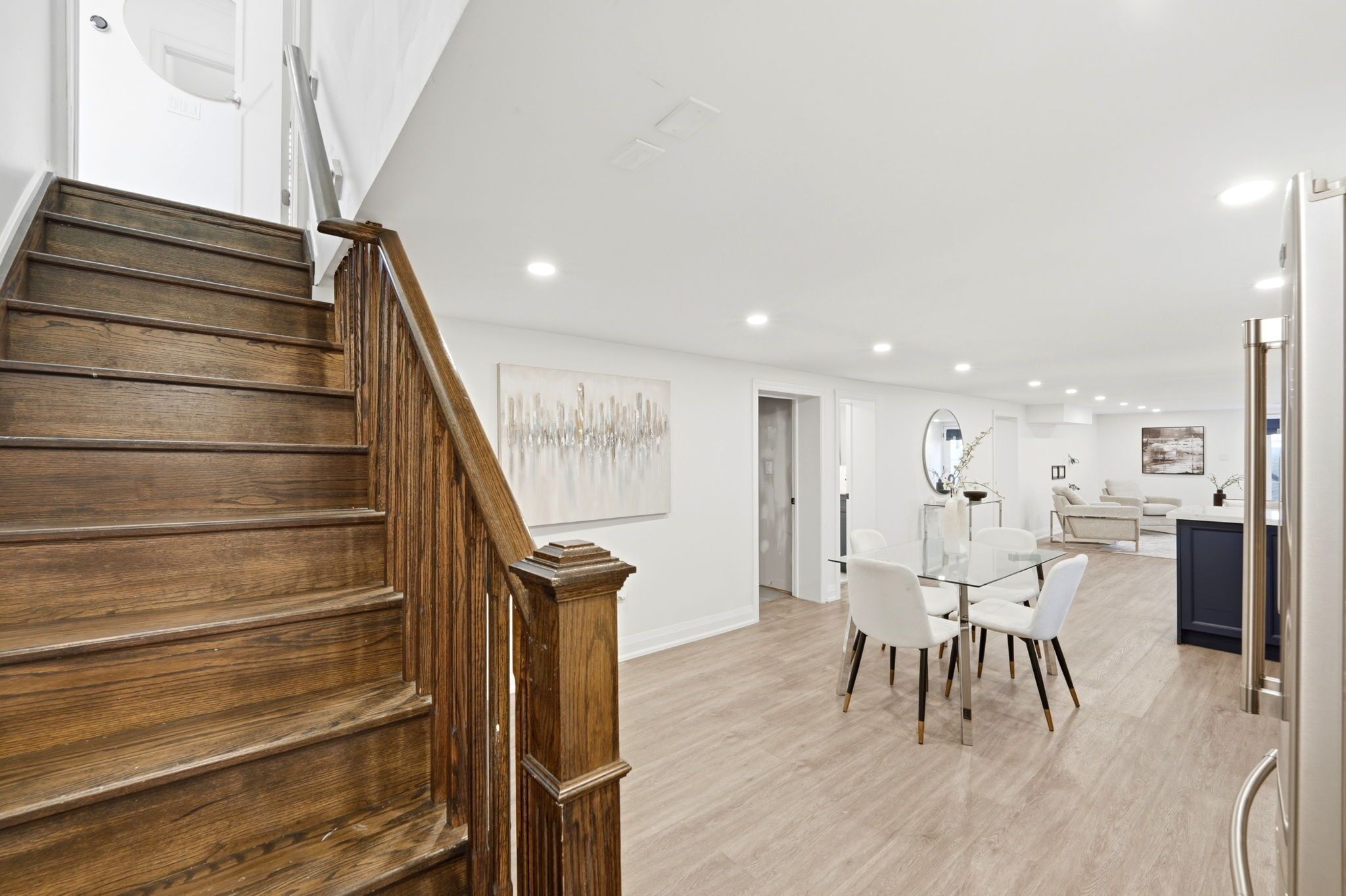 A modern interior with a wooden staircase, a dining area with white chairs, and a kitchen area.