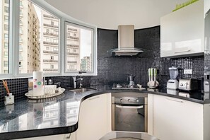 A modern kitchen with a large window, black tiled backsplash, stainless steel appliances, and a view of a residential building. - Step Into Elegance and Comfort (Santo Domingo)