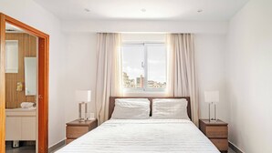 Ein Hotelzimmer mit einem großen Bett, zwei Nachttischen, einem Fenster mit Vorhängen und Blick auf Gebäude.