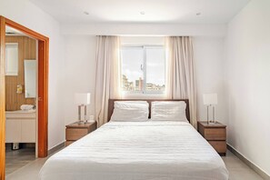 A hotel bedroom with a large bed, two bedside tables, a nightstand, a window with curtains, and a view of buildings. - Step Into Elegance and Comfort (Santo Domingo)