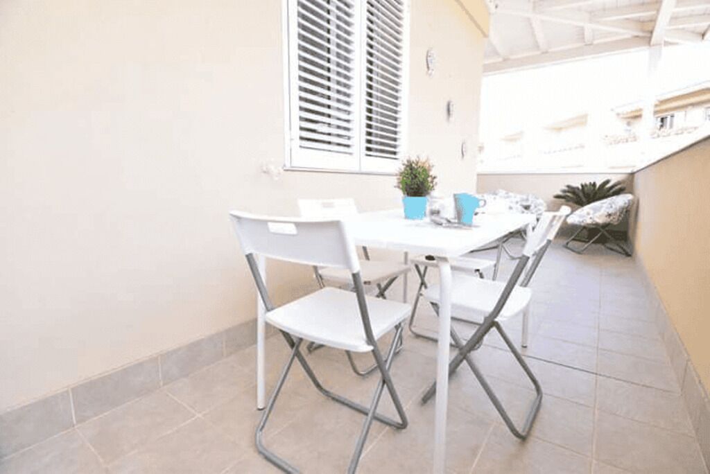 Un pequeño espacio exterior para comer con una mesa y sillas blancas, una planta en maceta y una ventana con persianas.