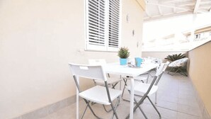 Uma área de refeições ao ar livre com uma mesa e cadeiras brancas, um vaso de planta e uma janela com persianas.