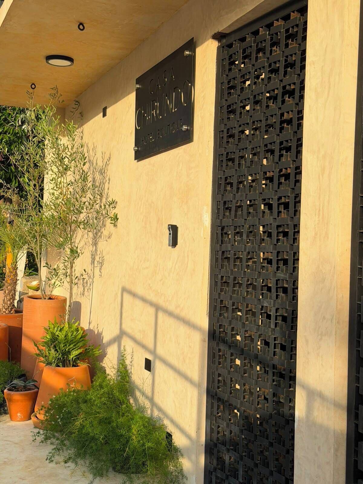 A building with a sign reading 'CASA CHIRINDO HOTEL ROUTE.' There are potted plants and a lattice screen on the facade.