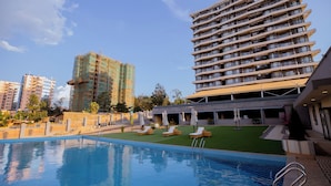 Una piscina con sillas de descanso y sombrillas, rodeada por un edificio con varios balcones y una obra en construcción al fondo.