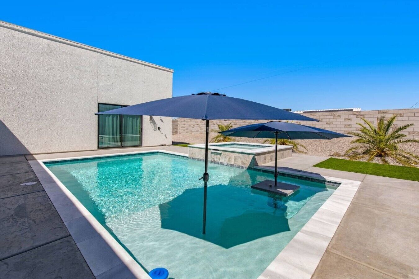 A rectangular swimming pool with a clear blue water surface, surrounded by a concrete deck. There are two umbrellas providing shade, and a small hot tub is visible in the background.