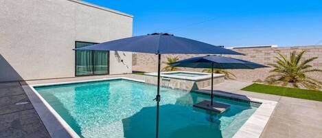 A rectangular swimming pool with a clear blue water surface, surrounded by a concrete deck. There are two umbrellas providing shade, and a small hot tub is visible in the background.