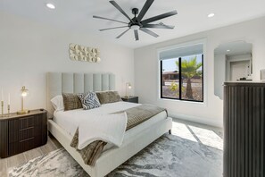 A bedroom with a bed, nightstands, a ceiling fan, a large window with a view, and a wall-mounted mirror. - Havasu Mcculloch - Luxury Feel, Luxury Stay (Lake Havasu City)