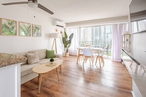 A modern living room with a sofa, coffee table, and dining area, featuring wooden flooring and large windows. - New Waikiki 2BR •Walk to Beach +free parking (Honolulu)