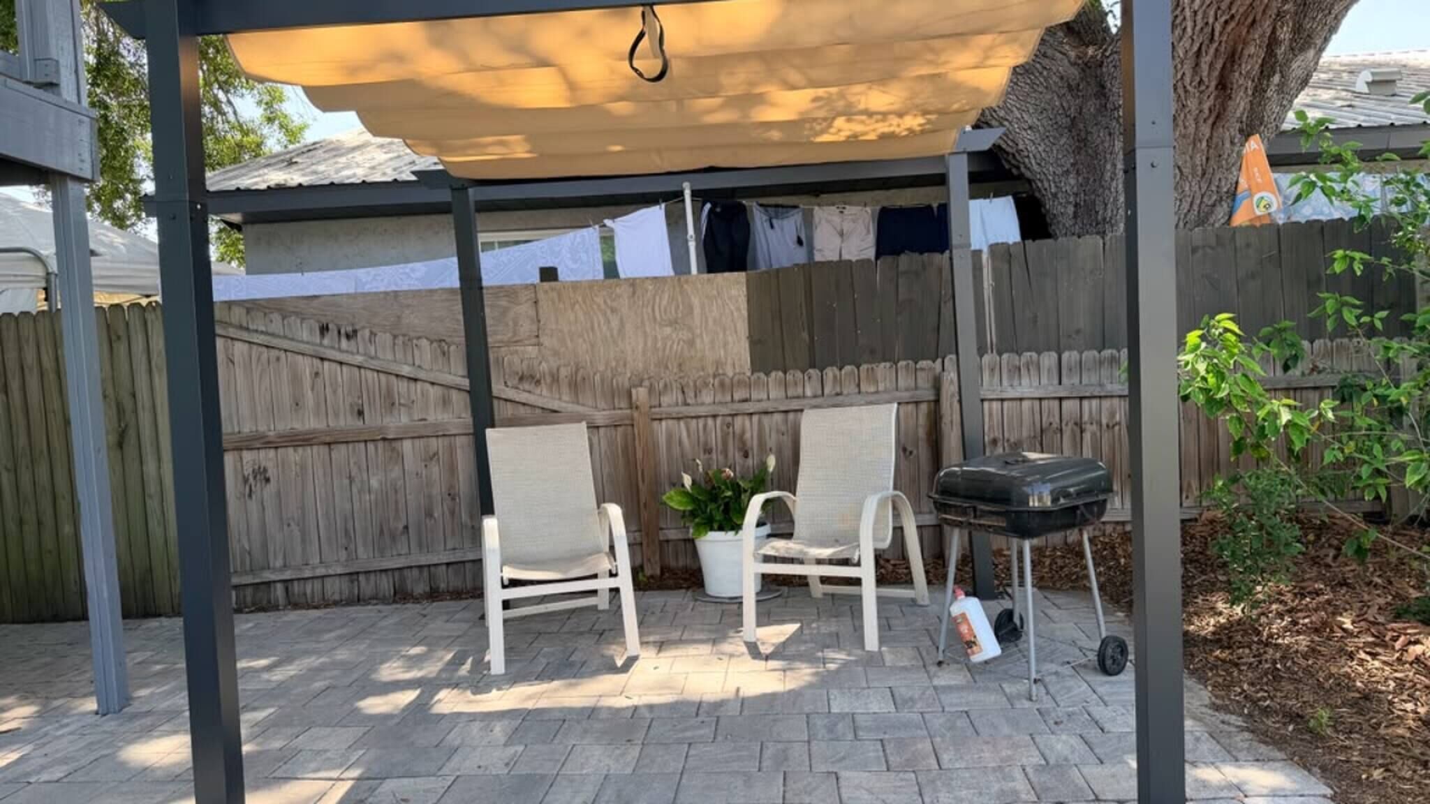 Ein überdachter Patio-Bereich mit Holzzaun, zwei weißen Stühlen, einem Grill und einem Topfpflanze.