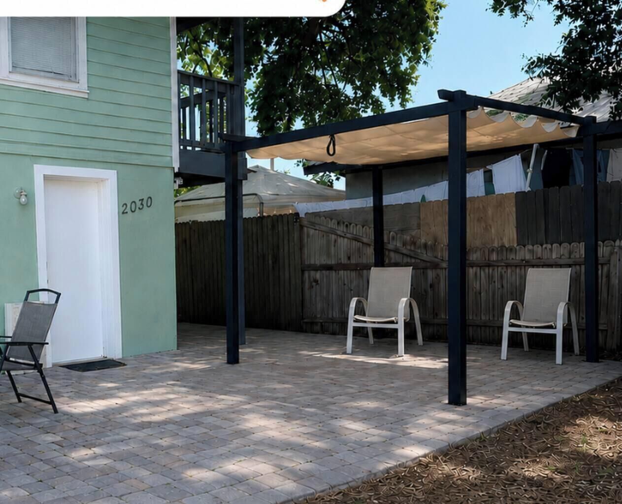 Een overdekt terrasgedeelte met twee stoelen en een tafel, een houten hek en het huisnummer 2030.