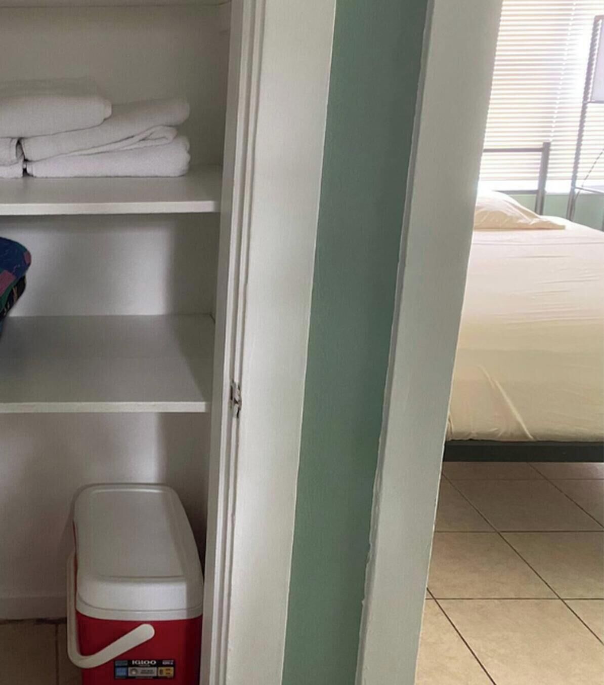 A hotel room with a bed, a shelf with folded towels, and a red cooler.